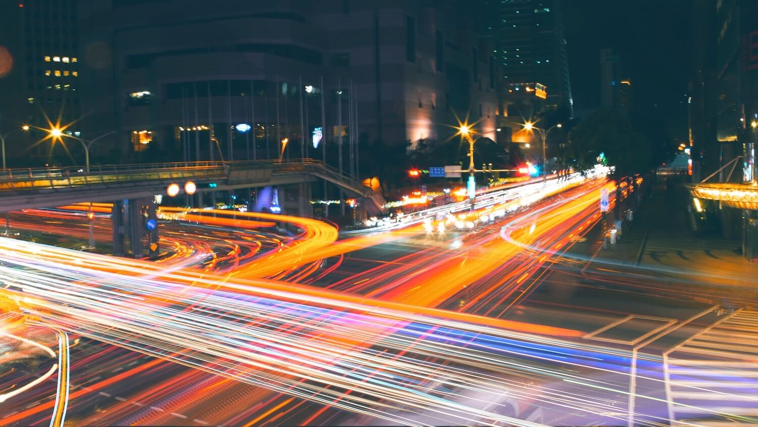 The blur of vehicle headlights and tail lights in a time-lapse photo of an intersection, city buildings visible in the background.