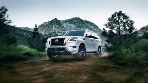 A white 2023 Nissan Armada full-size SUV driving on a dirt forest path with a snowy mountain in the background