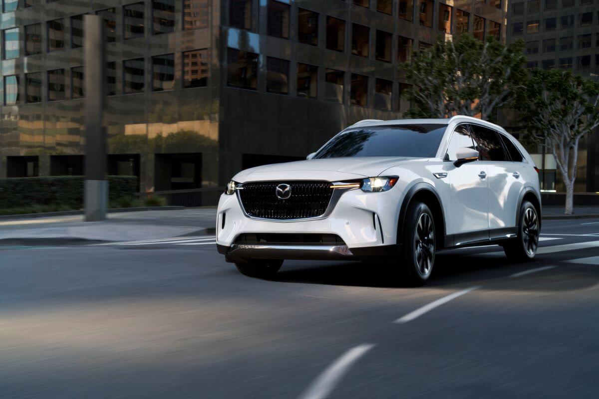 A white 2024 Mazda CX-90 full-size crossover SUV model driving through an urban city