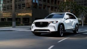 A white 2024 Mazda CX-90 full-size crossover SUV model driving through an urban city