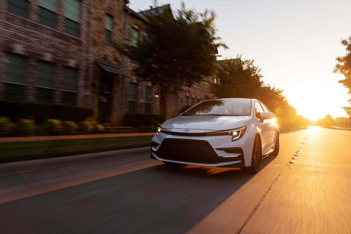 A 2023 Toyota Corolla XSE compact sedan model in Wind Chill Pearl driving past apartments as the sun sets. Toyota is one of the most reliable car brands.