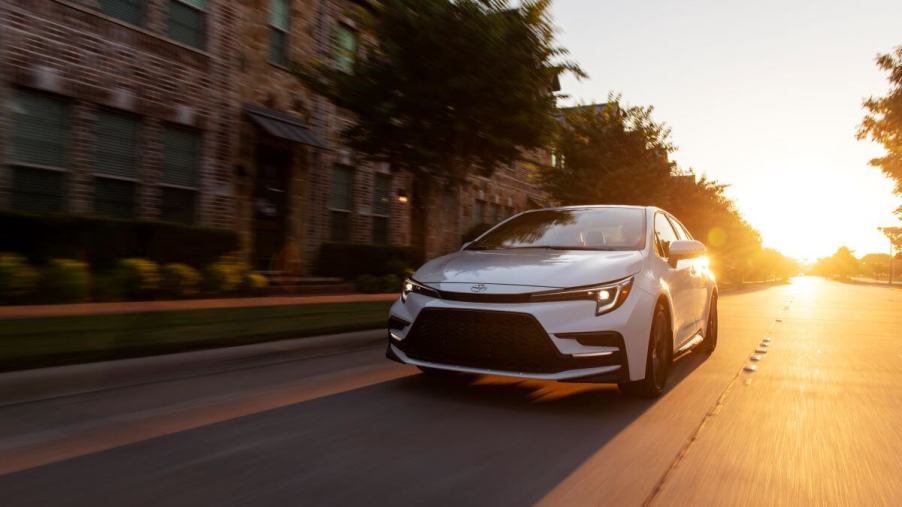 A 2023 Toyota Corolla XSE compact sedan model in Wind Chill Pearl driving past apartments as the sun sets. Toyota is one of the most reliable car brands.