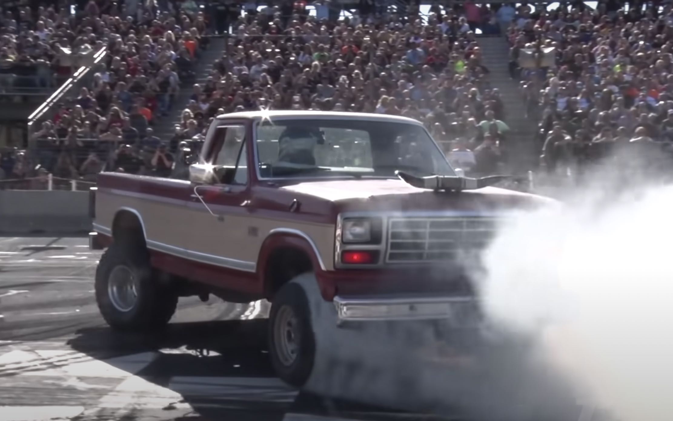 Smoke in front of a 1985 Ford F-150 truck modified to drive backward as it does burnouts, a crowd visible in the background.