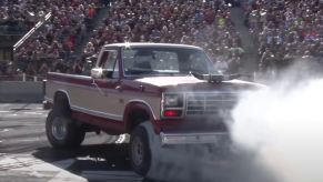 Smoke in front of a 1985 Ford F-150 truck modified to drive backward as it does burnouts, a crowd visible in the background.