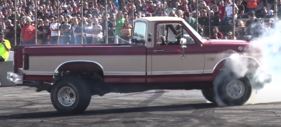 A modified 1985 Ford F-150 that can drive backwards does a burnout at the Indianapolis Speedway, a crowd visible in the background.