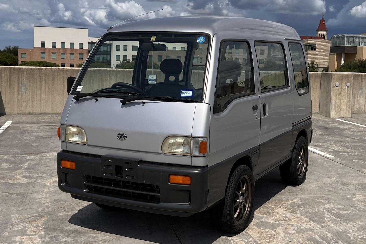 A silver 1995 Subaru Sambar mini bus parked at the top of a parking garage, a city skyline in the background.