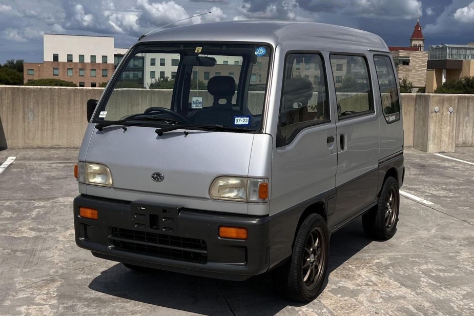 A silver 1995 Subaru Sambar mini bus parked at the top of a parking garage, a city skyline in the background.