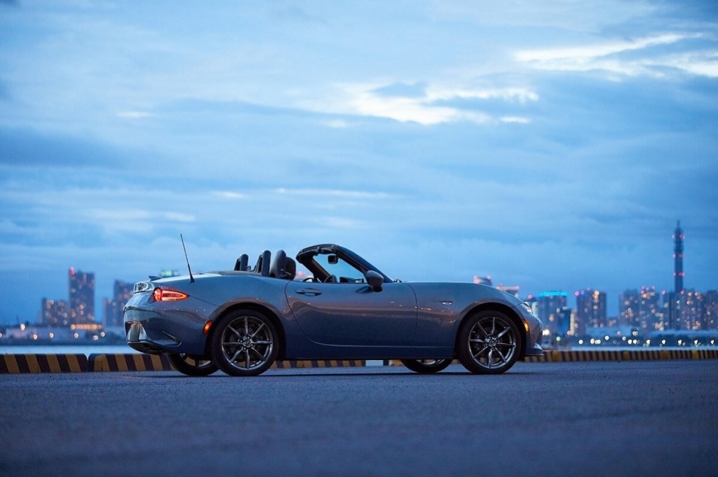 A gray Mazda MX-5 Miata convertible sports car parks next to a city skyline. 