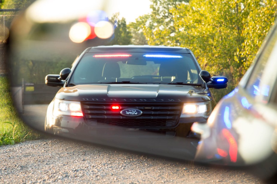 A rearview mirror framing an unmarked Ford Interceptor police car with its lights on, parked in front of trees.