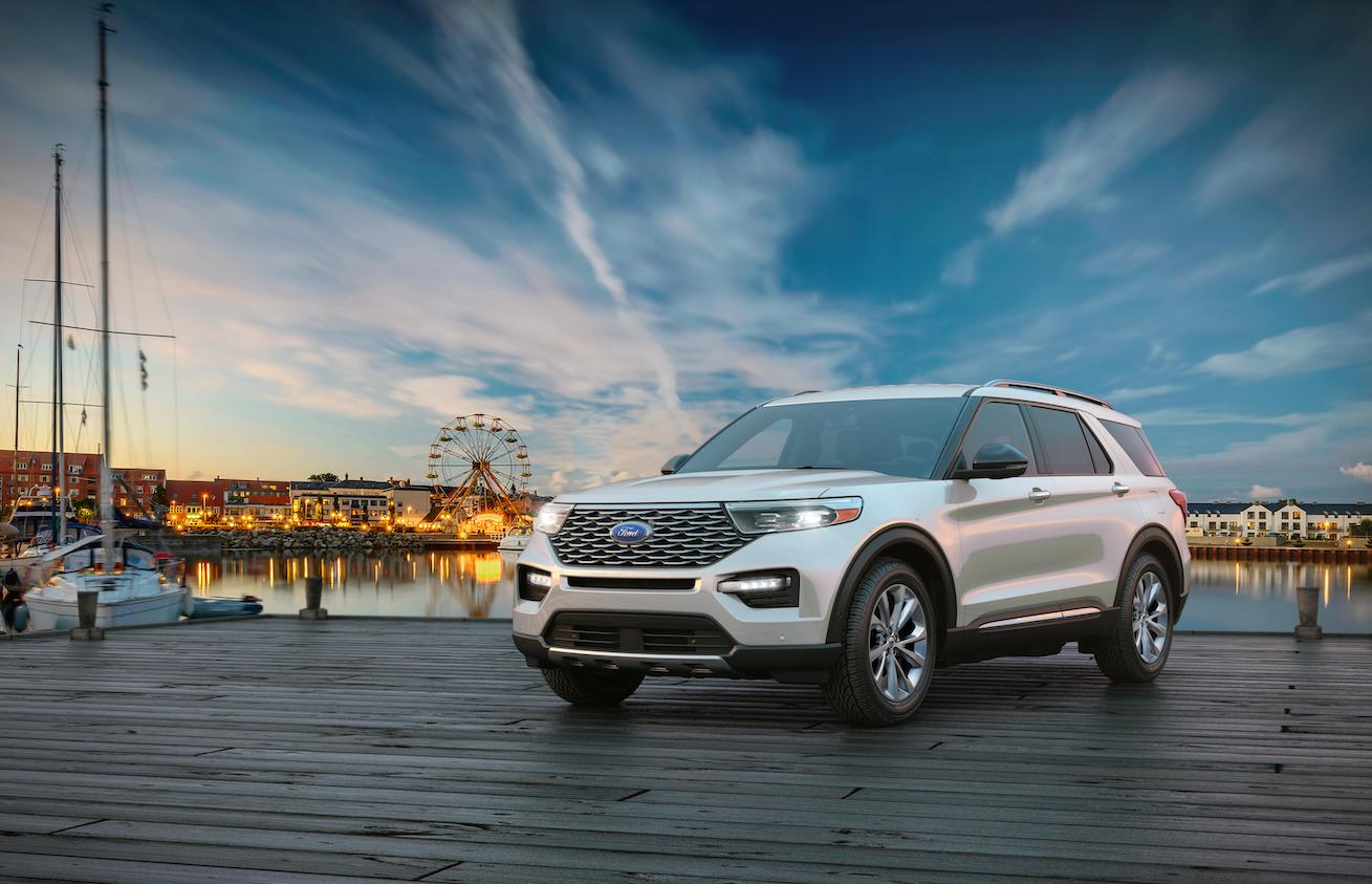 A white 2023 Ford Explorer parked on a pier with a carnival behind it. Ford sales include favorable numbers for the Explorer.