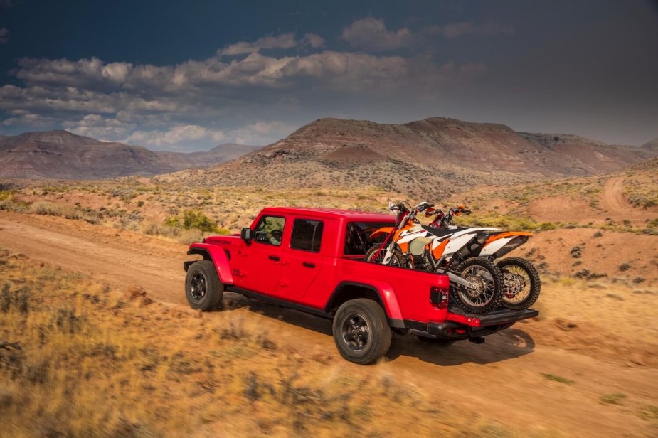 A red Jeep Gladiator pickup truck carries two dirt bikes on a 4WD trail, desert mountains visible in the background.