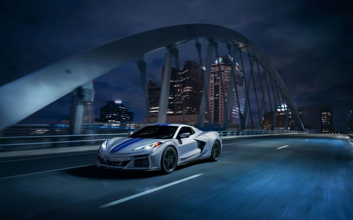 A silver 2024 Chevy Corvette E-Ray 3LZ convertible with blue racing stripes crossing a bridge at night