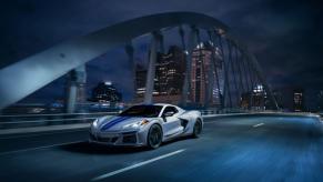A silver 2024 Chevy Corvette E-Ray 3LZ convertible with blue racing stripes crossing a bridge at night