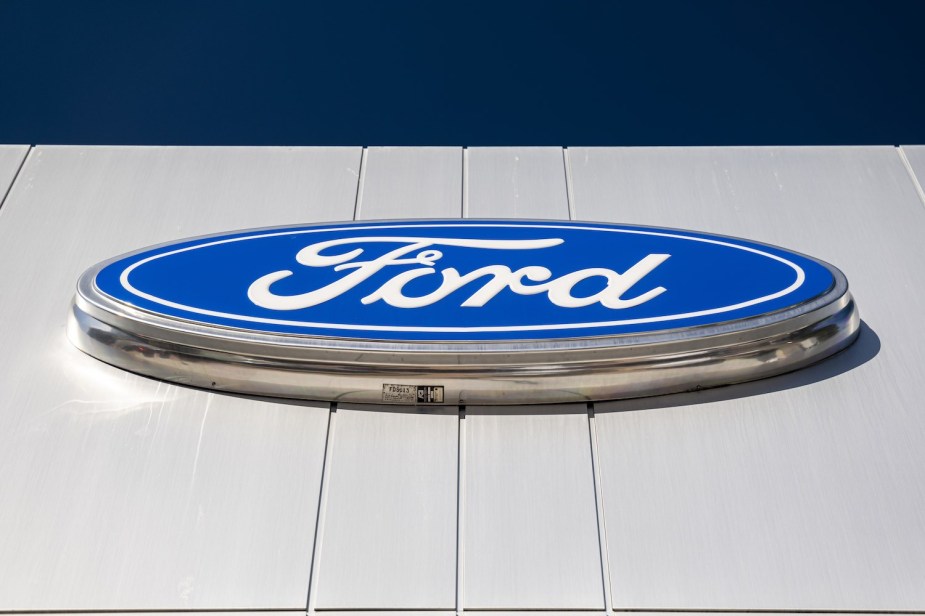 The Ford logo on the wall of a dealership that sells many configurations of the F-150 truck, blue sky visible in the background.