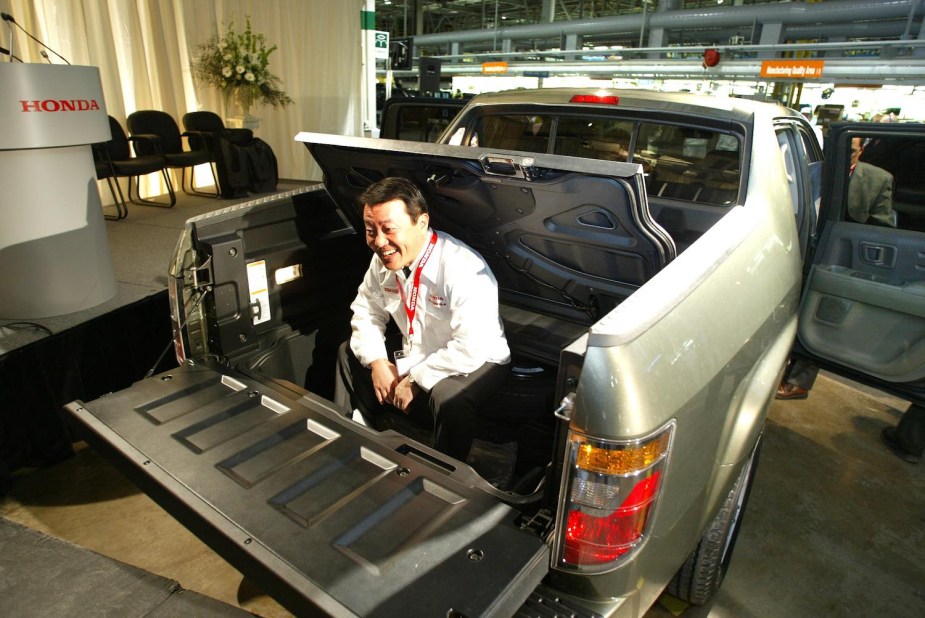 The CEO of Honda Canada poses in the in-bed trunk of the first Ridgeline compact truck during a press conference, a stage visible in the background.