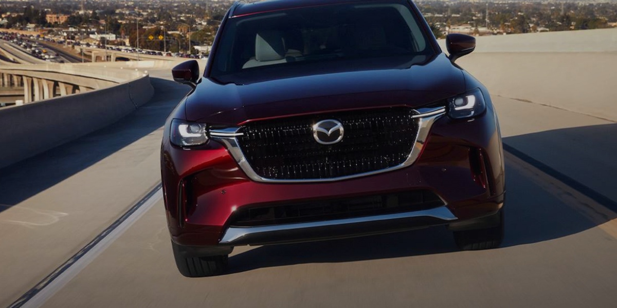 A red 2024 Mazda CX-90 midsize three-row SUV is driving on the road.