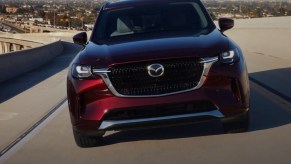 A red 2024 Mazda CX-90 midsize three-row SUV is driving on the road.