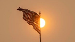 A U.S.A. flag waves in front of a hazy, setting sun like the flags in front of auto plants in the U.S.