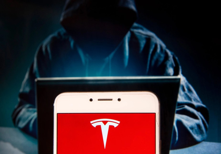 A hacker behind a Tesla logo using a laptop with a hood on. 