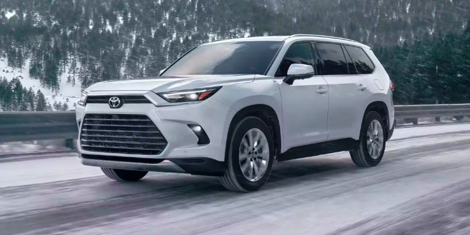 A white 2024 Toyota Grand Highlander midsize SUV is driving on a snowy road. 