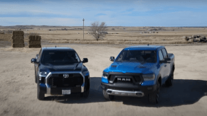 A dark blue Toyota Tundra and light blue Ram 1500 parked next to one another on a farm, hay bales in the background.