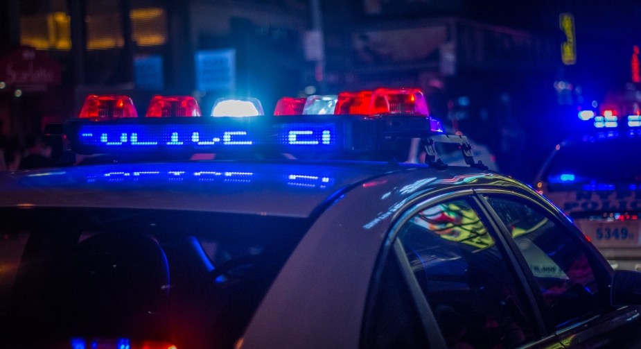 The red and blue light bar on the roof of a marked police car keeping a crime scene safe for traffic, more cars visible in the distance.