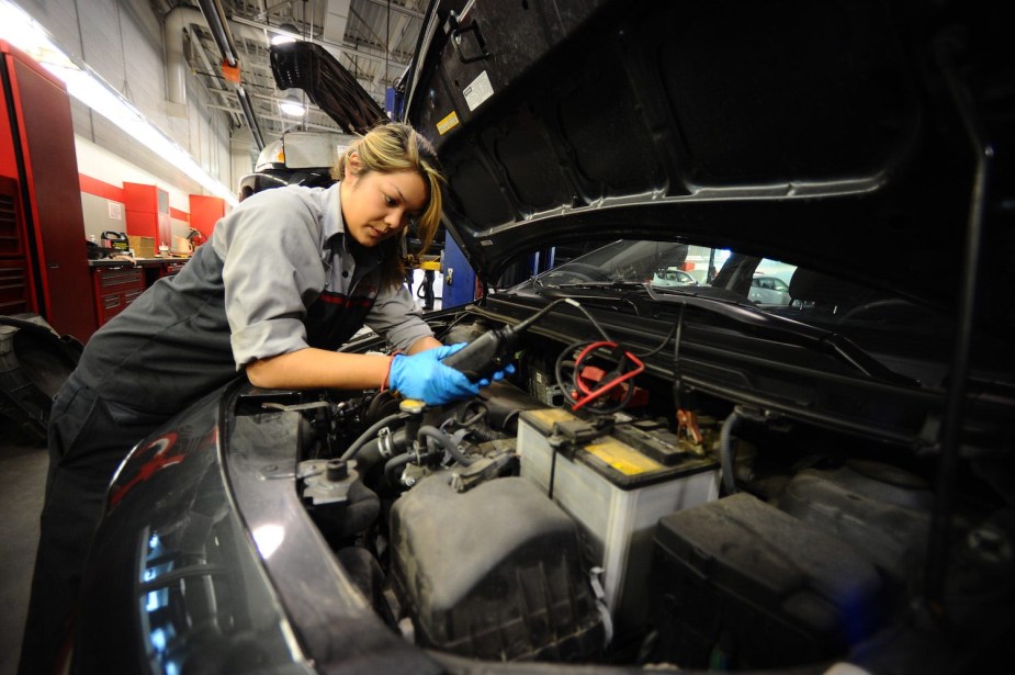 An automotive service technician connects a sensor to the battery of a minivan, the shop visible behind her.