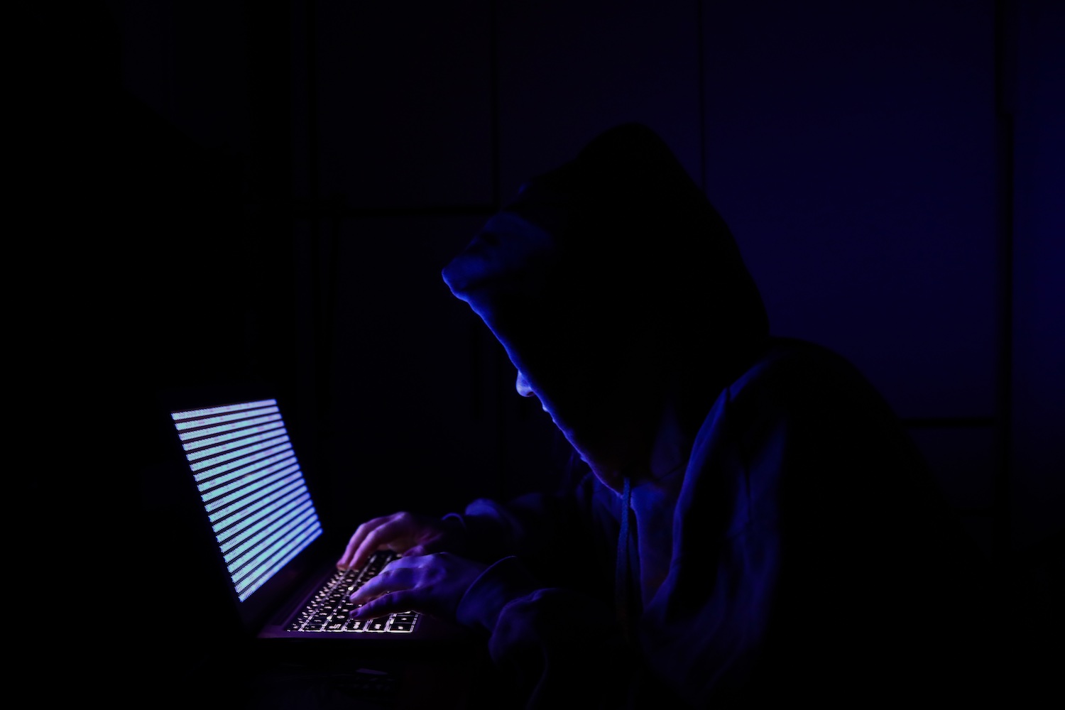 A hacker in a hoodie sweatshirt hunched over a laptop.