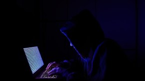 A hacker in a hoodie sweatshirt hunched over a laptop.