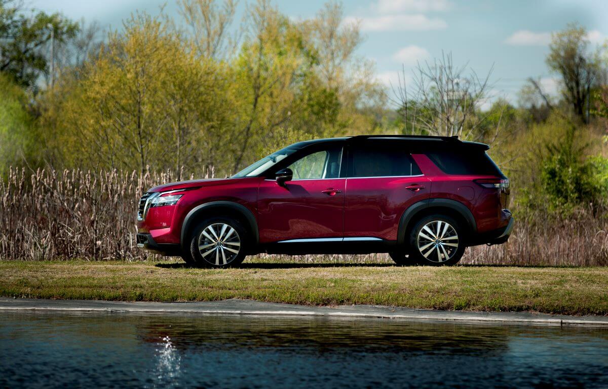 A side profile shot of a 2023 Nissan Pathfinder midsize SUV model parked on a grass shore near a small lake or pond
