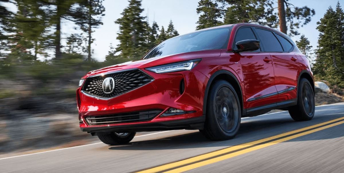 A 2024 Acura MDX midsize luxury crossover SUV model in red driving on a forest country highway