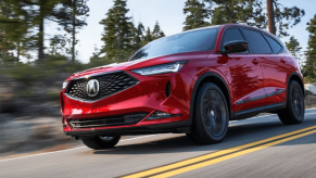 A 2024 Acura MDX midsize luxury crossover SUV model in red driving on a forest country highway