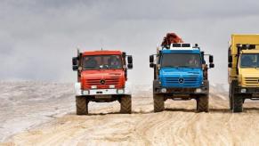 A line of Mercedes-Benz Unimog models in red, blue, and yellow driving down a road in a desert