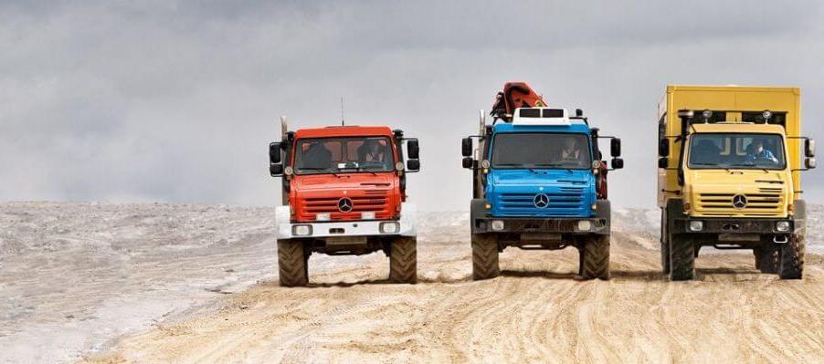 A line of Mercedes-Benz Unimog models in red, blue, and yellow driving down a road in a desert