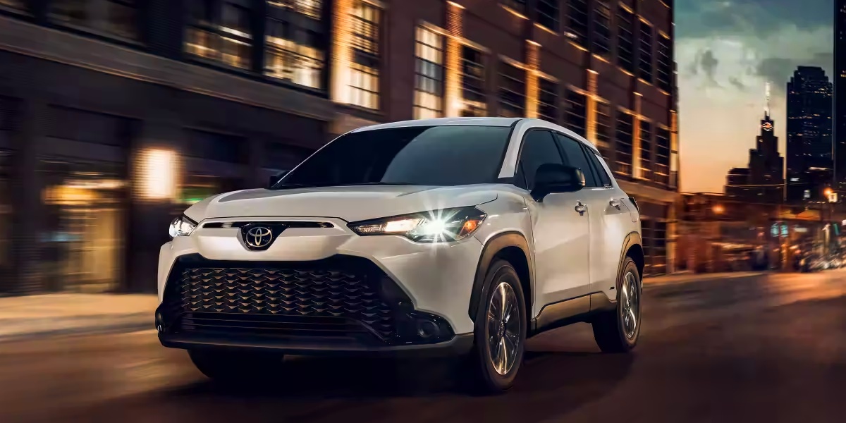 A white 2023 Toyota Corolla Cross Hybrid subcompact hybrid SUV is driving on the road.