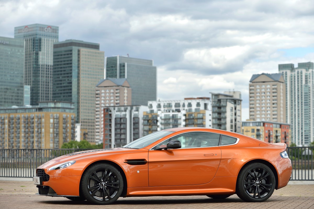A used Aston Martin Vantage in orange, parked in front of a cityscape.