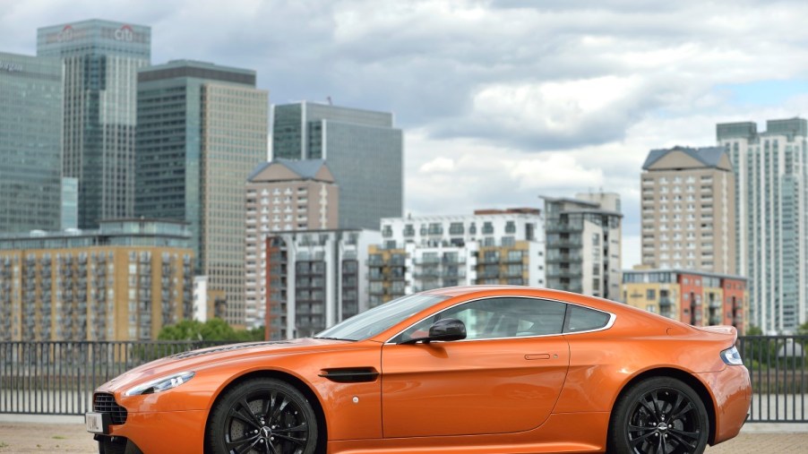 A used Aston Martin Vantage in orange, parked in front of a cityscape.