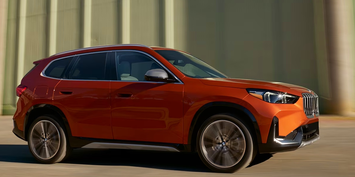 An orange 2023 BMW X1 luxury subcompact SUV is driving on the road.