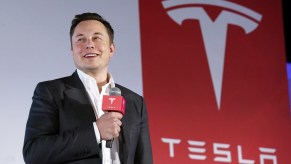 Elon Musk speaks next to his electric car company's emblem, a Tesla "T" logo.