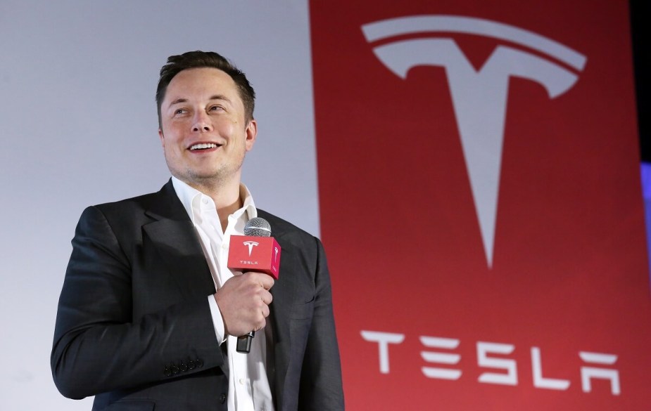 Elon Musk speaks next to his electric car company's emblem, a Tesla "T" logo.
