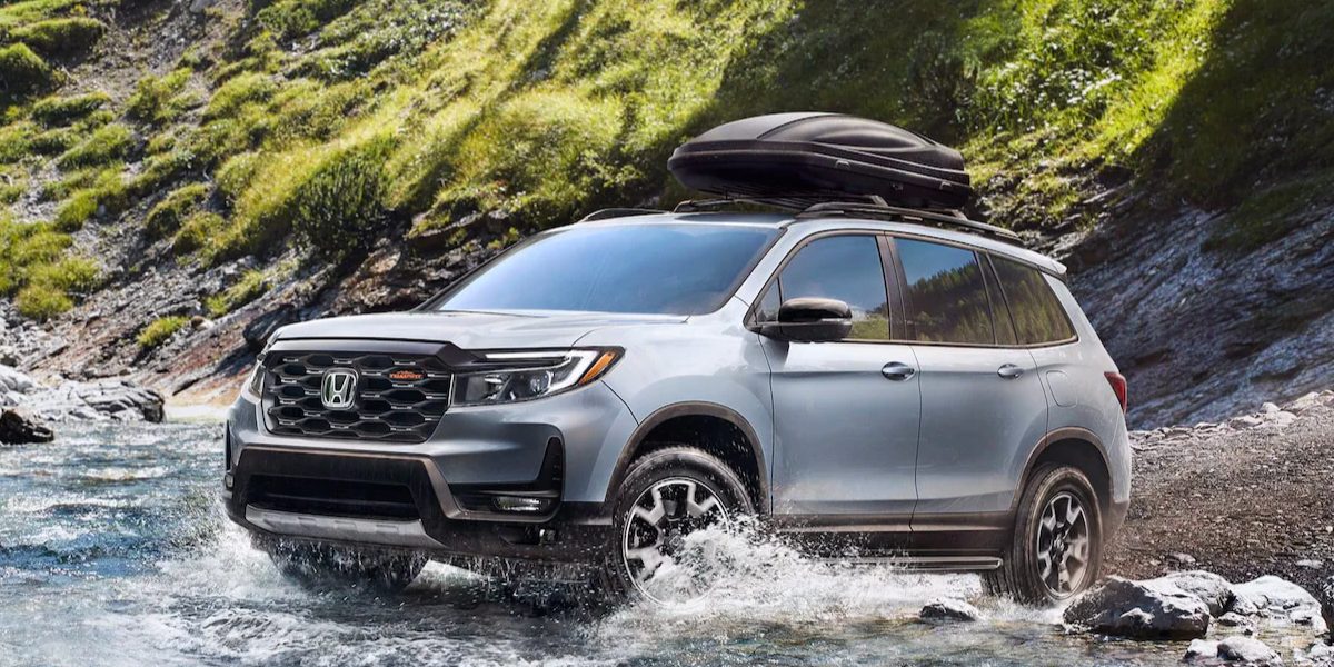 A gray 2023 Honda Passport midsize SUV is driving through shallow water off-road.