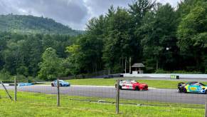 An Aston Martin, BMW, and Porsche GT4 racecars ascend the hill at Lime Rock Park