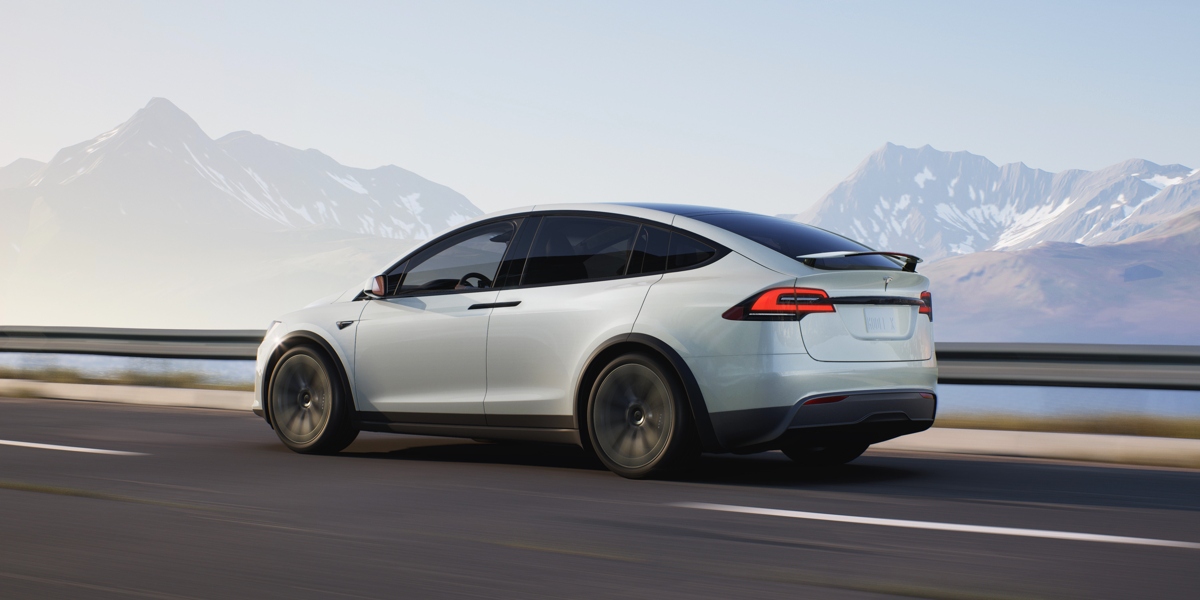 A white 2023 Tesla Model X midsize SUV is driving on the road.