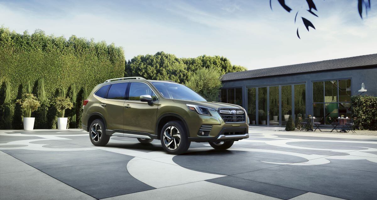 A light-green 2023 Subaru Forester compact SUV model parked on a open plaza surrounded by tall hedges