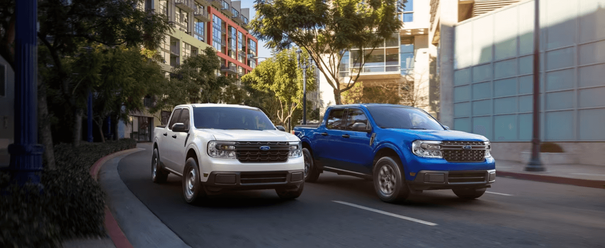 White and blue 2024 Ford Maverick compact pickup truck models driving under tree shade in a city downtown
