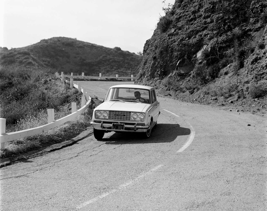 White 1966 Toyota Corona cornering on a steep road, mountains visible in the background.