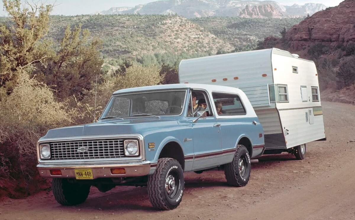 A pale-blue 1970 Chevy Blazer towing a travel trailer on a dirt road