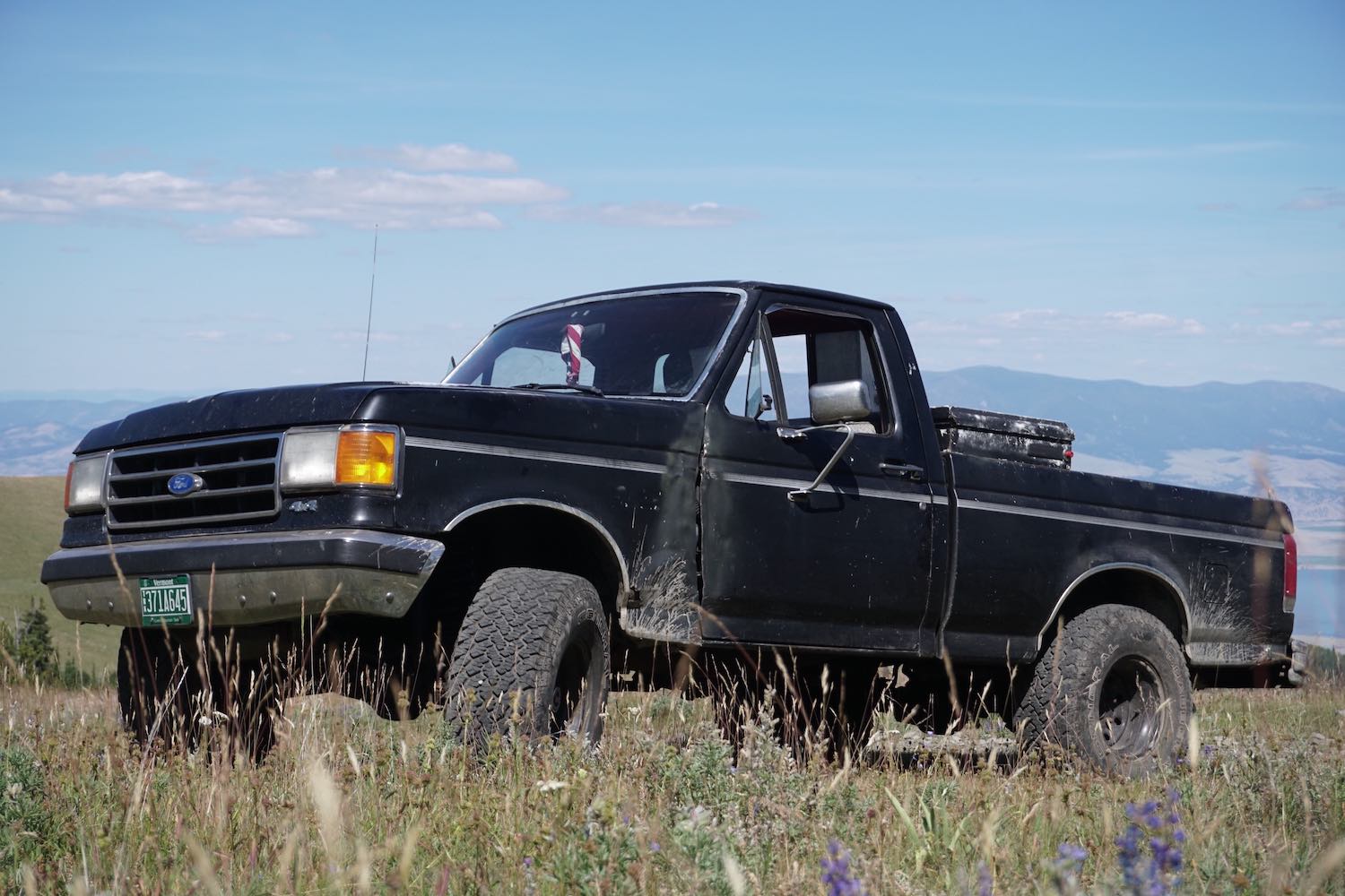 Black square body Ford F-150 classic pickup truck parked in a field, blue sky visible in the background.