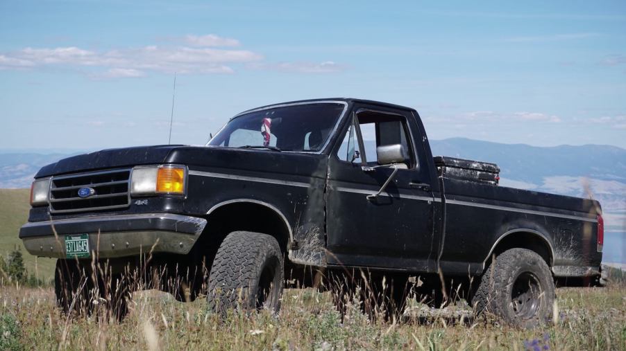 Black square body Ford F-150 classic pickup truck parked in a field, blue sky visible in the background.