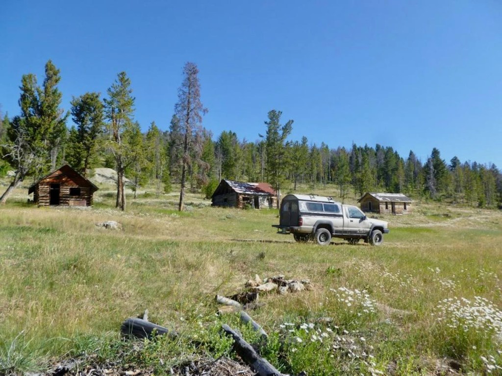 A tan Mitsubishi pickup truck with a camper parked in front of a ring of abandoned cabins at a Wyoming ghost town.